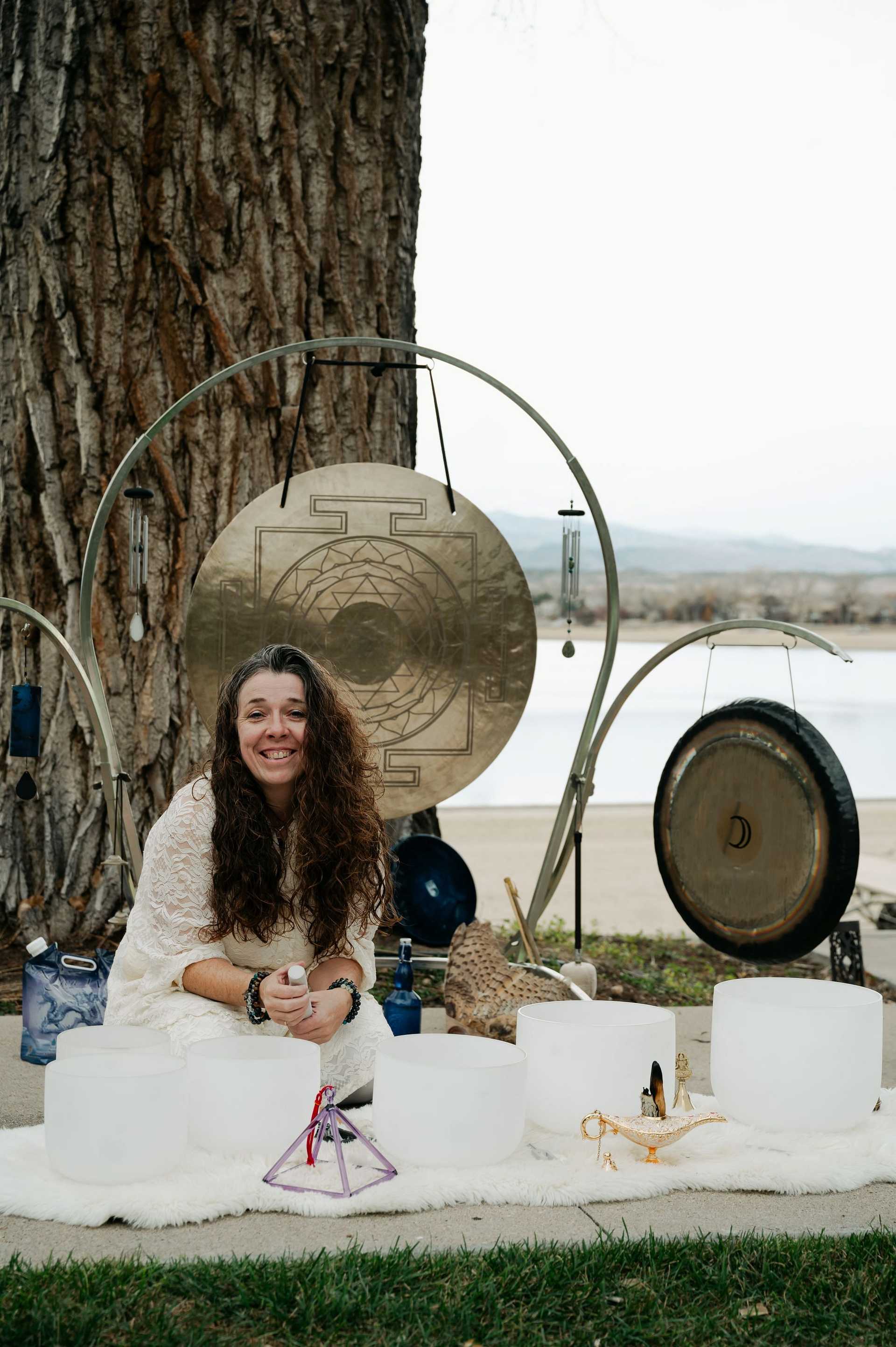 Meditative setup with a smiling person, sound bowls, gongs, and instruments by a tree outdoors.