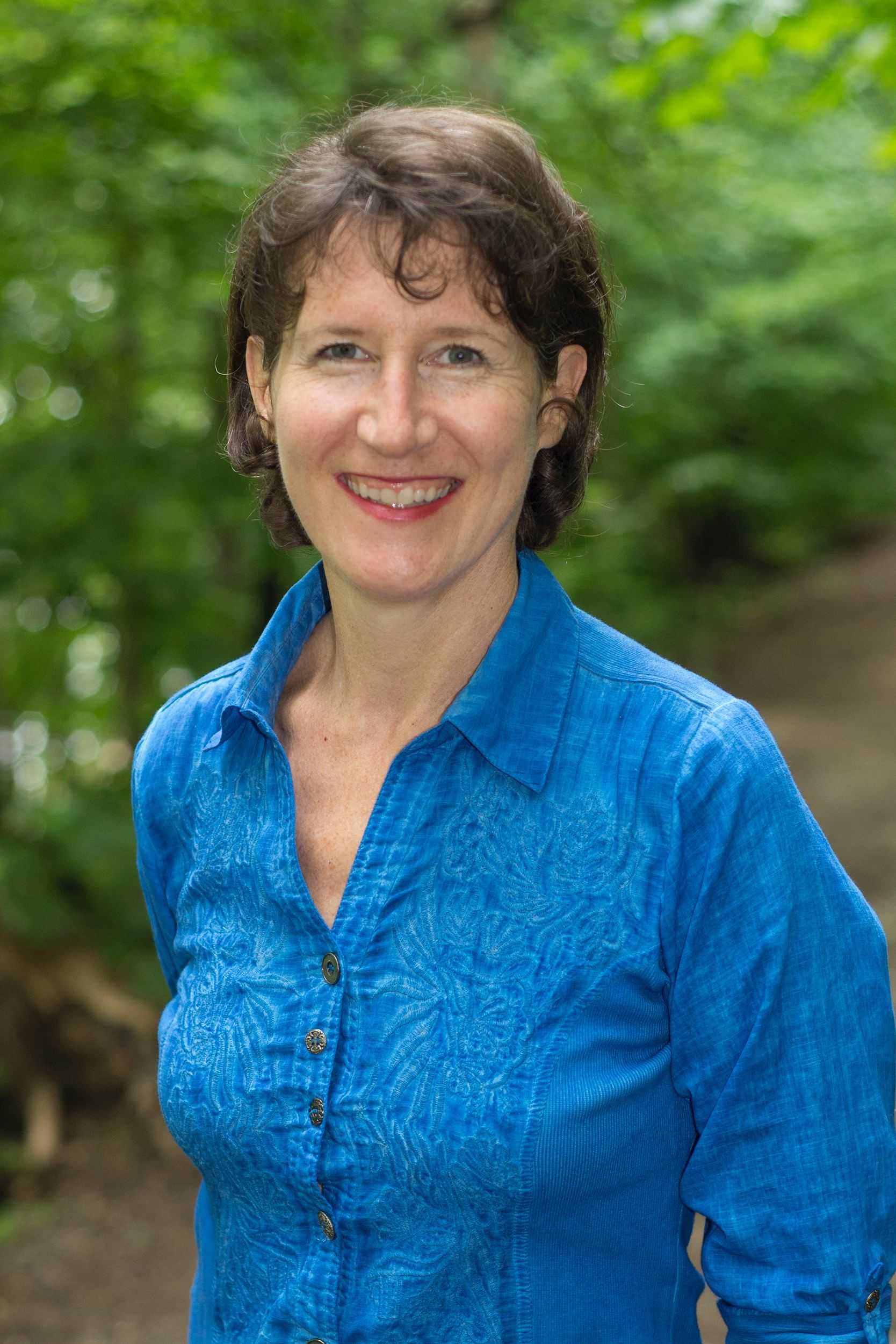 Smiling woman in a blue shirt standing on a forest path.